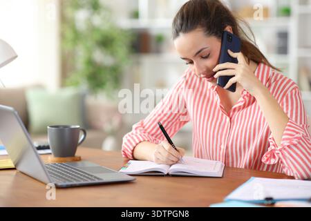 Freelance worker writing in agenda talking on phone working at home Stock Photo