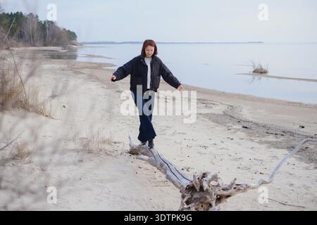 Woman balancing on driftwood log at sandy beach. Concept of life balance, mindfulness, solitude and outdoor activity in autumn nature. Stock Photo