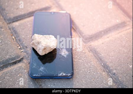 A broken smartphone with a cracked screen lies on the paving slabs; nearby lies the stone that caused the damage. Stock Photo
