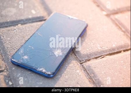 A smartphone with a broken screen lies on the tiled sidewalk: cracks and chips are visible in the glass, the casing is scratched, and the photo was ta Stock Photo