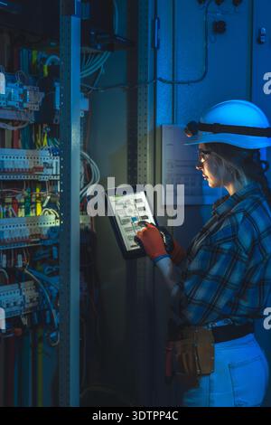 Female electrician reviews control cabinet wiring on tablet in plant. Side profile check. Stock Photo