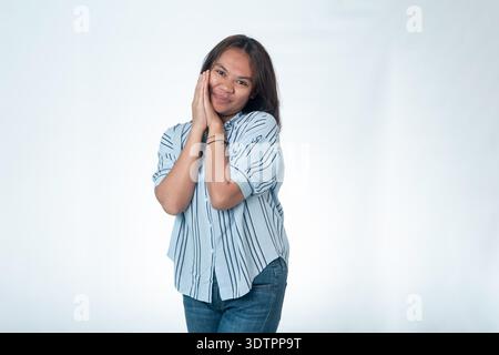 Portrait of a charming Indonesian woman in a blue striped shirt leaning her cheek against her folded hands. She has a shy, happy, and blushing express Stock Photo