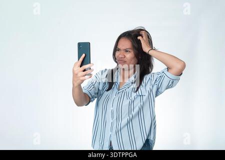 Indonesian woman in a blue striped shirt looking at her smartphone with an annoyed and confused expression while scratching her head. Isolated on whit Stock Photo