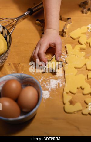 A child's hand is using a star-shaped cookie cutter on yellow pastry dough, prepared on a wooden surface. Flour is sprinkled around the dough. A metal Stock Photo