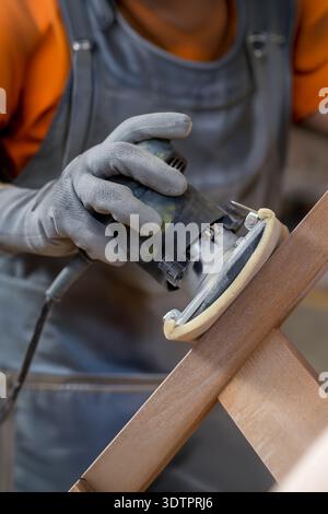 In a carpentry workshop, a worker is sanding a wooden stool using an electric sander. The person, wearing gloves and a work apron, focuses on smoothin Stock Photo