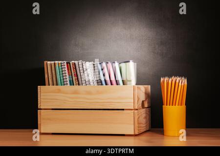 Back to school, On table there is wooden box filled with notebooks and glass with pencils against  background of black school board, chalk board Stock Photo