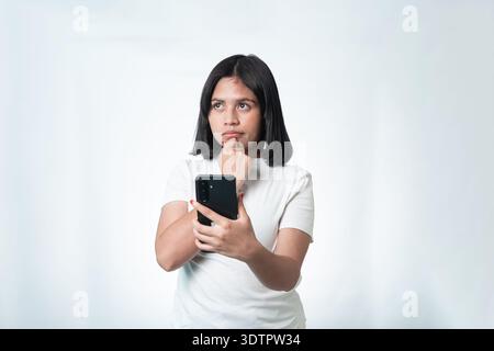 Young Indonesian woman in a white t-shirt holding a mobile phone and looking away with a pensive or confused expression. Isolated on white studio back Stock Photo