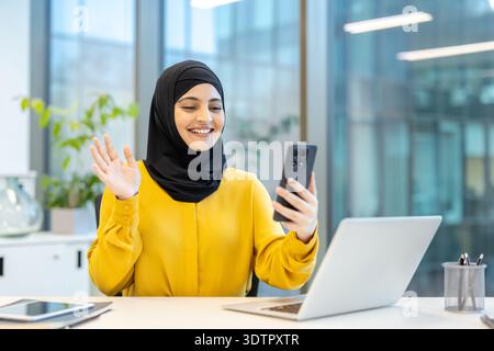 Muslim woman in hijab smiling and waving during a video call on her smartphone while working at an office desk with laptop, showing friendly remote communication and professionalism Stock Photo