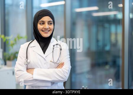 Young smiling muslim female doctor standing with crossed arms in a hospital hallway, wearing a white lab coat and a stethoscope, representing professionalism and diversity in healthcare Stock Photo