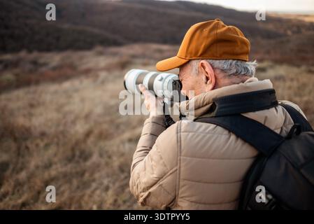 A man wearing a cap, padded jacket, and backpack standing in mountain, focusing through his camera lens to photograph something. Stock Photo