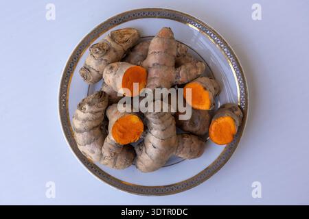 Top view of fresh raw turmeric (Curcuma longa) roots and slices on a white plate. Organic rhizomes showing bright orange inside. Isolated on white bac Stock Photo