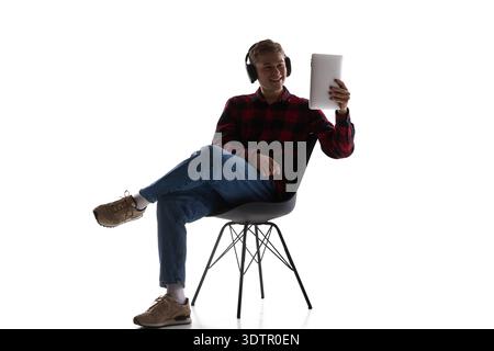 Young man wearing headphones using tablet sitting on chair isolated on white. Stock Photo