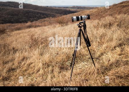 Digital camera with large telephoto lens standing on tripod in dry grass landscape on the mountain. Stock Photo