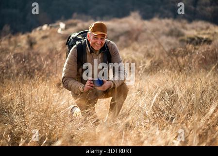 A nature photographer kneels in a field of dry grass, carefully adjusting his camera settings while capturing close-up details of the landscape in war Stock Photo