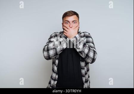 A young man stands with his hands covering his mouth, eyes wide with shock and astonishment. He wears a stylish checkered shirt and conveys strong emo Stock Photo