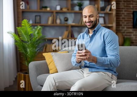 Young man sitting on a sofa in his modern living room, smiling and absorbed in his smartphone while browsing apps, social media, streaming or messaging for leisure and connection Stock Photo