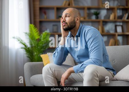 Thoughtful bald man sitting alone on a gray sofa in a modern living room, looking away while pondering, reflecting, and experiencing stress or disappointment Stock Photo