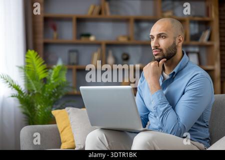 Young adult man on a sofa with a laptop, looking away while contemplating ideas, problem-solving, and strategizing for his remote work from a comfortable home office environment Stock Photo