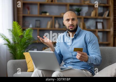 Young man sitting on a sofa, looking confused and shrugging while holding a credit card and using a laptop, experiencing difficulties with online banking or shopping Stock Photo
