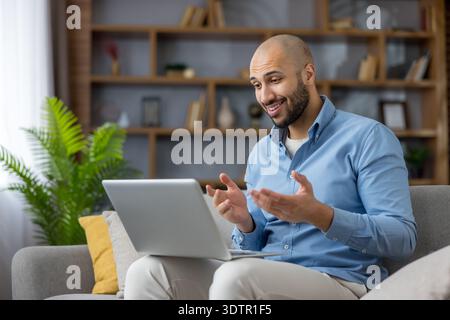 Smiling african american man on a sofa in a modern home office, confidently engaging in a laptop video call for remote work, presentation or virtual meeting, gesturing while speaking Stock Photo