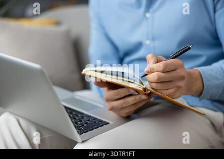 Man holding an open notebook and writing notes with a pen while sitting with a laptop on his lap, focusing on work, planning, and remote learning concepts Stock Photo