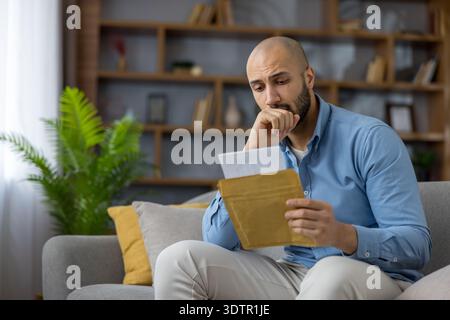 Stressed bald man receiving disappointing financial news, holding a brown envelope and document with a concerned expression while sitting on a sofa in his living room Stock Photo