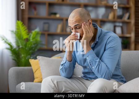 Young man at home feeling unwell, blowing his nose into a tissue while suffering cold or flu symptoms congestion, headache, fatigue and seasonal allergy discomfort Stock Photo