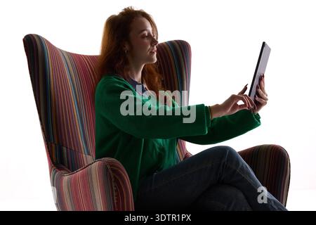 Young woman using tablet while sitting in striped armchair isolated on white. Stock Photo