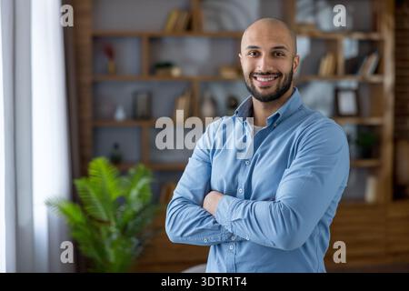 Confident bald businessman with beard and crossed arms, smiling in a modern office workspace with bookshelves behind him, projecting leadership, success and approachability Stock Photo