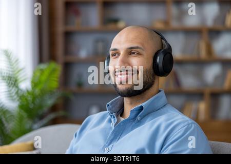 Young man wearing a blue shirt and black wireless headphones, smiling happily while relaxing and enjoying digital audio in a comfortable home environment Stock Photo
