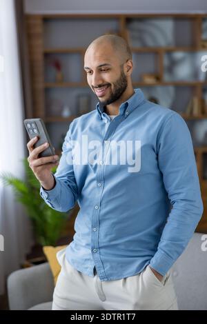 Smiling man enjoying a digital connection, holding a smartphone while interacting with online content or messages, representing modern technology and communication in a casual home setting Stock Photo