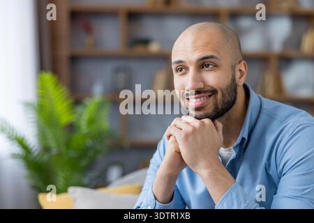 Content bald man in a blue shirt smiling and looking away, daydreaming and contemplating future plans in a relaxed, comfortable home setting with warm, hopeful expression Stock Photo