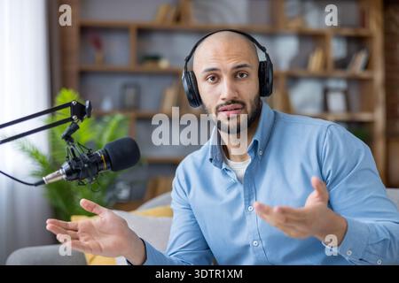 Young man wearing headphones and speaking into a microphone, recording an online podcast or streaming from his home studio, actively communicating with listeners Stock Photo