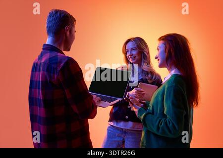 Three young professionals discussing project using laptop and tablet on gradient background. Stock Photo