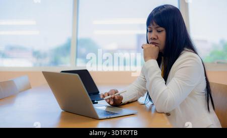 Asian businesswoman working on laptop and tablet in office. Female professional thinking, analyzing data, using stylus pen for digital tasks. Professi Stock Photo