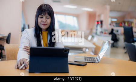 Happy Asian businesswoman working with digital tablet and laptop in modern office. Professional female entrepreneur using wireless technology for onli Stock Photo