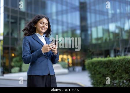 Young businesswoman standing outdoors in front of a modern glass building, actively using her smartphone and smiling, embodying communication and professional connectivity Stock Photo