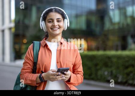Young student woman standing outdoors with white headphones and a backpack, using her smartphone and smiling at the camera, conveying modern education and connectivity Stock Photo