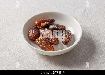 A small white bowl filled with fresh, dark brown dates resting on a textured gray surface. Stock Photo