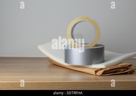Two rolls of tape, one clear and one metallic, resting on a wooden table with a folded paper and a white cloth in the background. Stock Photo