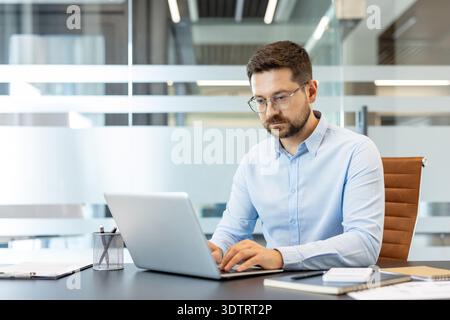 Young businessman wearing glasses and a light blue shirt concentrating on his laptop while typing at a desk in a contemporary corporate office environment Stock Photo