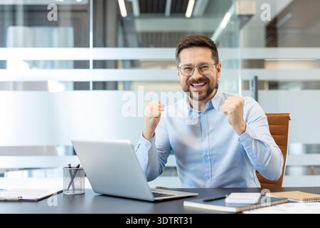 Happy businessman feeling successful and celebrating achievement with clenched fists while smiling broadly, sitting at a modern office desk with a laptop Stock Photo