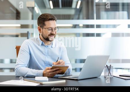 Young businessman wearing glasses sitting at a desk, looking at his laptop screen and writing diligent notes in a spiral notebook, showcasing productivity in a modern office environment Stock Photo