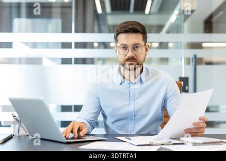 Serious businessman working in a bright modern office, holding documents in one hand and typing on a laptop with the other, processing information and managing business tasks Stock Photo