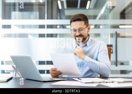 Professional man wearing glasses reviewing financial papers, calculating data or studying business reports while working at desk with laptop in corporate workspace Stock Photo