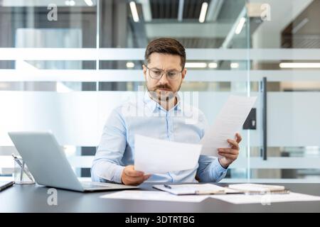 Businessman in glasses carefully reviewing financial documents and contracts, analyzing data and concentrating on important paperwork while working at a modern office desk Stock Photo