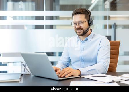 Young man wearing a headset with a microphone and light blue shirt sitting at a modern office desk, typing on a laptop, providing customer service and support assistance Stock Photo