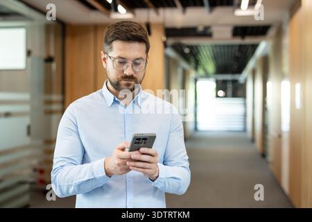 Businessman wearing eyeglasses and a blue shirt holding mobile phone, texting, and browsing in a modern office hallway, representing remote communication and connectivity Stock Photo
