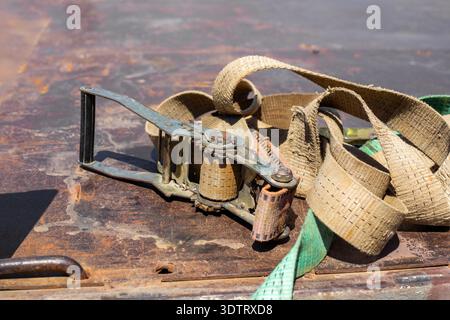 A dirty beige ratchet strap lies on a rusty metal surface after securing a load. industrial equipment, cargo safety, transport tie down. Stock Photo