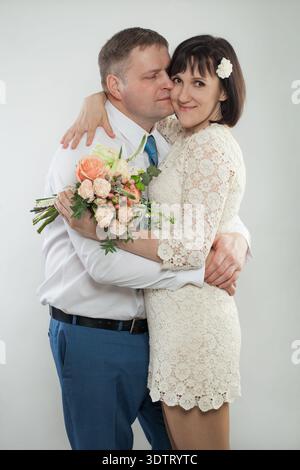 A couple stands close together, sharing a joyful moment on their wedding day. The man smiles while the woman holds a bouquet of flowers. They appear h Stock Photo